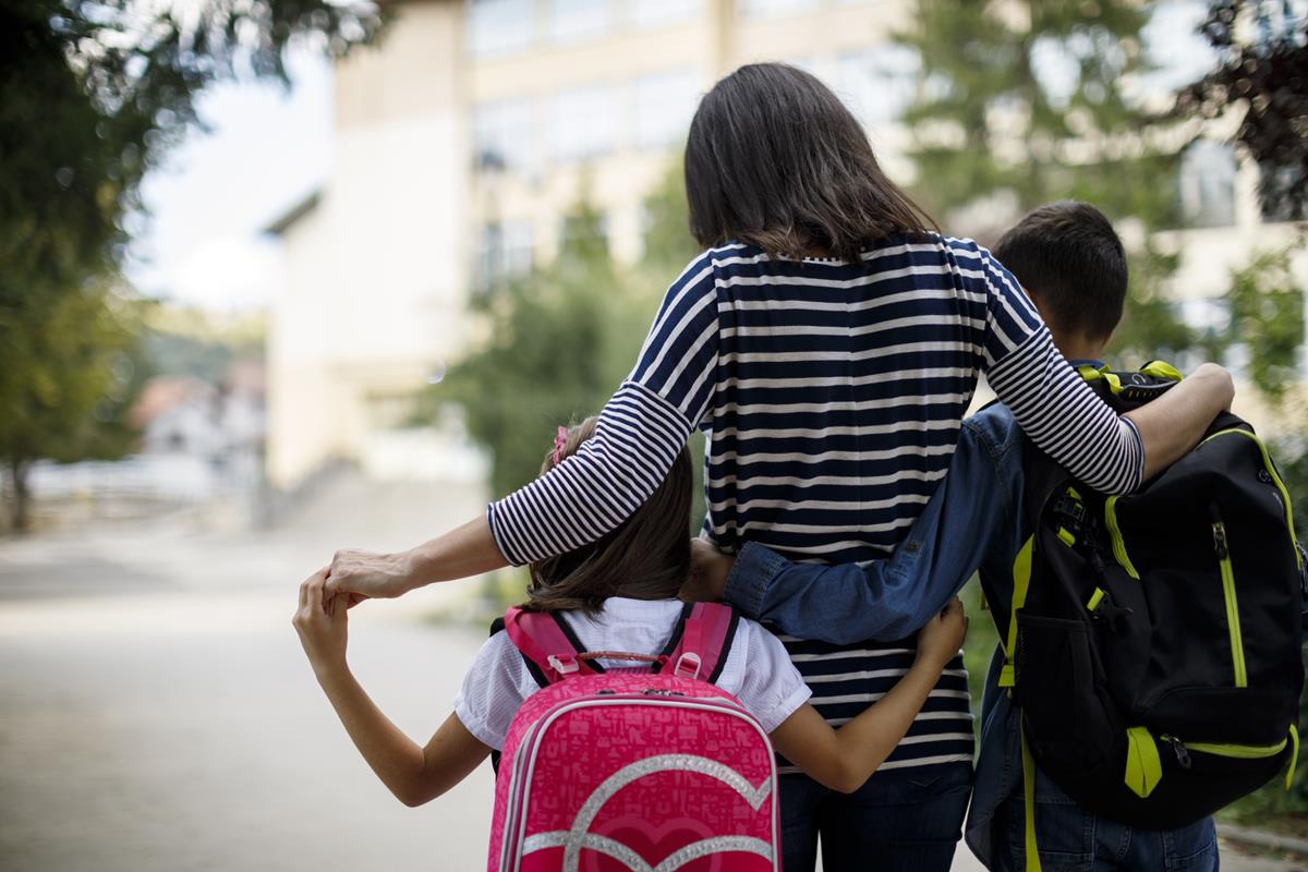 Two elementary aged children walking on either side of an older woman, likely their caretaker, with their arms wrapped around each other. They are walking away from the camera.